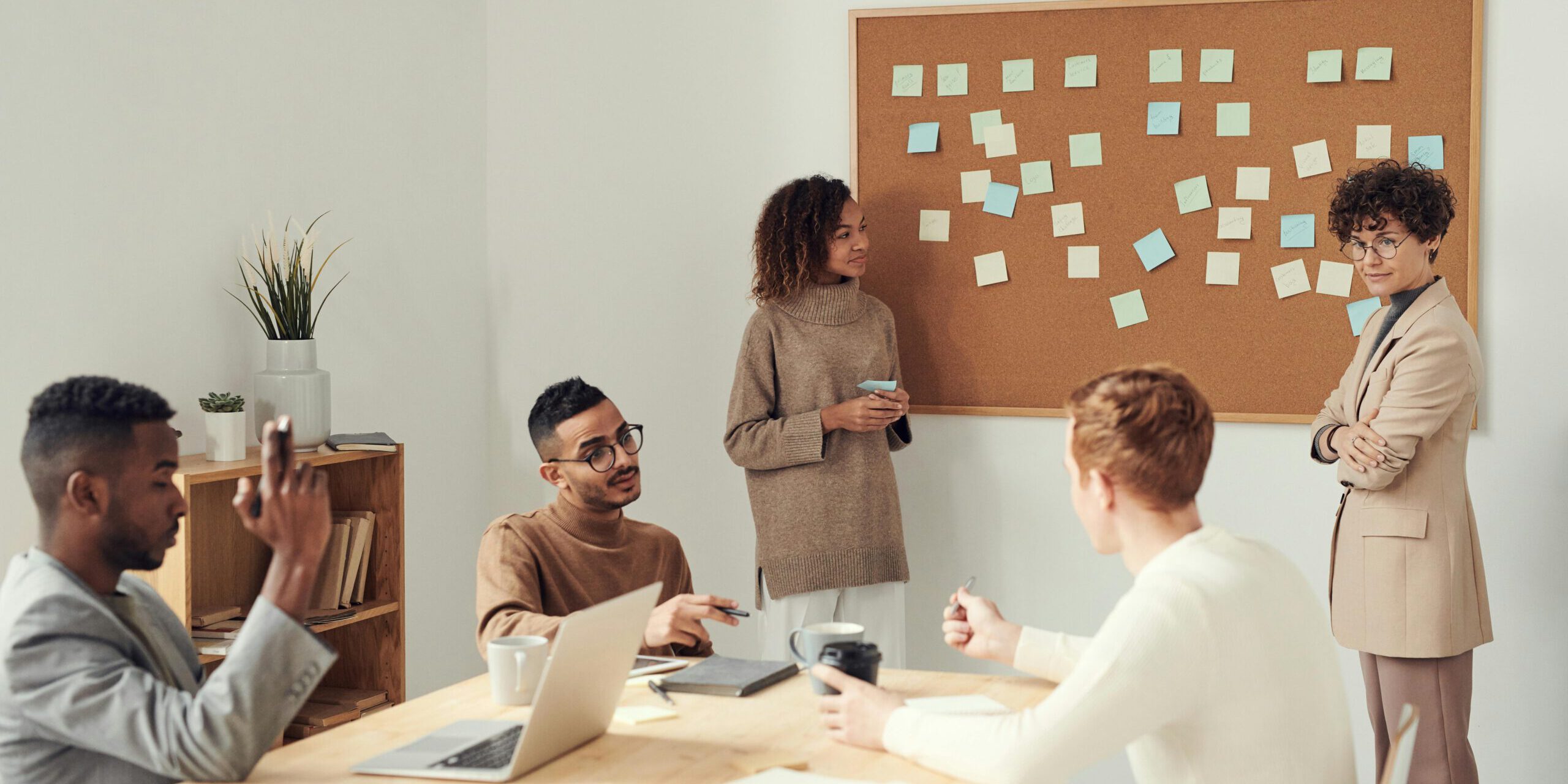 vijf professionals zitten en staan rondom een tafel terwijl ze in overleg zijn. Er staat een laptop op tafel en twee professionals zijn bezig met een groot bord met post-its