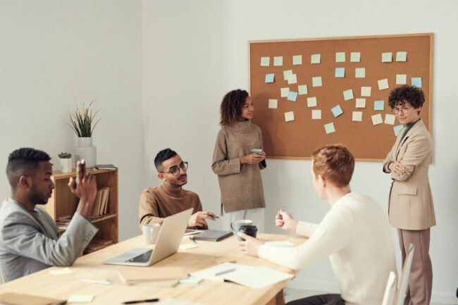 vijf professionals zitten en staan rondom een tafel terwijl ze in overleg zijn. Er staat een laptop op tafel en twee professionals zijn bezig met een groot bord met post-its